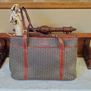 May include: A rectangular tote bag with a geometric pattern in shades of gray and brown. The bag features brown leather accents, including the handles and trim. A small statue and a wooden decorative piece are in the background.
