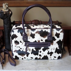 Könnte beinhalten: Rechteckige Handtasche mit Kuhfellmuster, weißem Grund und schwarzen und braunen Flecken. Sie hat lila Griffe, einen Reißverschluss und einen abnehmbaren Schultergurt. Die Tasche hat auch eine Reißverschlusstasche vorne.