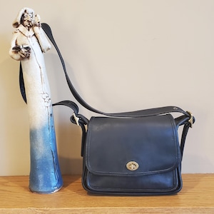 May include: A black leather shoulder bag with a fold-over flap, a gold-toned metal turn-lock closure, and an adjustable shoulder strap. The bag is positioned next to a tall, slender ceramic sculpture of a figure with a white and blue finish.