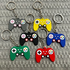 May include: Seven colourful video game controller keyrings with silver key rings. The controllers are in shades of grey, black, green, red, blue, and yellow.