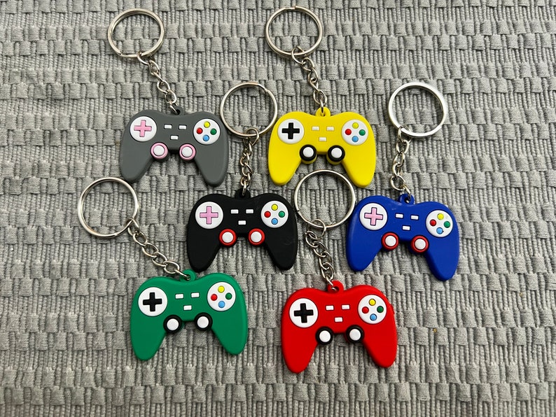Puede incluir: Siete llaveros de mando de videojuegos coloridos con anillas de plata. Los mandos son en tonos de gris, negro, verde, rojo, azul y amarillo.