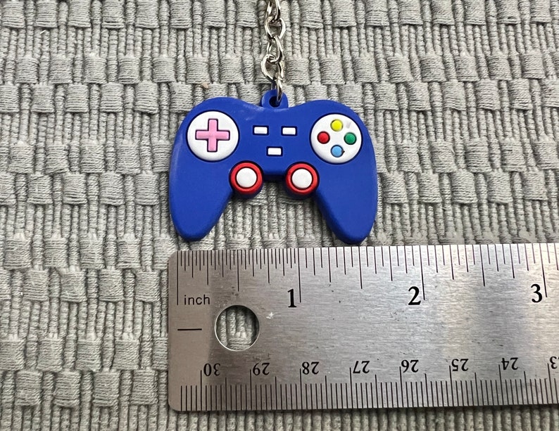 Puede incluir: Llavero de pl&aacute;stico azul en forma de mando de videojuego con un dise&ntilde;o de botones rosa, amarillo, verde y azul. El llavero mide aproximadamente 2,5 cm de largo.