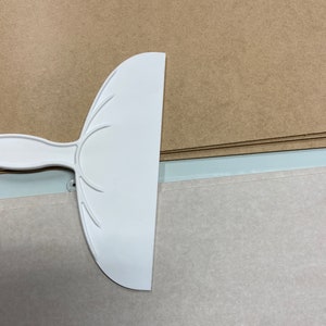 May include: White plastic scraper tool with a curved blade and a handle. The scraper is on a brown surface.