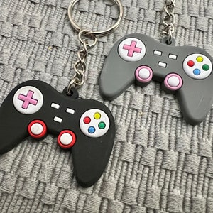 Puede incluir: Dos llaveros de pl&aacute;stico de color negro y gris con forma de mando de videojuego. Un llavero es negro con botones rojos y blancos y una almohadilla de botones multicolor. El otro llavero es gris con botones rosas y blancos y una almohadilla de botones multicolor.