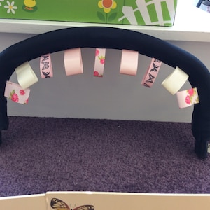 May include: A black stroller handle with a variety of pink and white ribbons tied around it. The ribbons have floral and butterfly patterns.