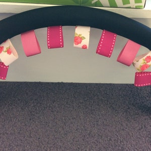 May include: Black stroller handle with pink and white ribbon loops attached. The ribbons have a floral pattern and white polka dots.
