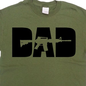 May include: Olive green t-shirt with the word "DAD" in large black letters. The letter "A" is replaced with a black silhouette of a rifle. The shirt is made of a soft material and has short sleeves.
