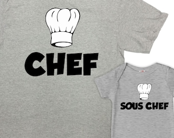 Könnte beinhalten: Ein graues T-Shirt mit einer Kochmütze und dem Wort "CHEF" darauf gedruckt. Ein kleinerer grauer Baby-Strampler mit einer Kochmütze und dem Wort "SOUS CHEF" darauf gedruckt.