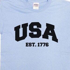 May include: Light blue t-shirt with the word "USA" in bold black letters and "EST. 1776" underneath. The shirt is made of a soft material and has short sleeves and a crew neck.