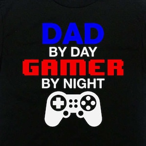 Puede incluir: Camiseta negra con el texto "Dad by day Gamer by night" en letras azules y rojas. Un gráfico de mando de videojuego blanco está debajo del texto.