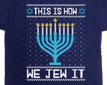 May include: Navy blue t-shirt with a festive design. The shirt features a blue menorah with lit candles and the text "THIS IS HOW WE JEW IT" in white. The design has a pixelated, sweater-like appearance.