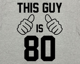 Puede incluir: Camiseta gris con texto negro que dice "This Guy Is 80" con dos pulgares hacia arriba. El texto está en negrita, en tipo de letra de bloque. Hay una pegatina roja en forma de estrella con el texto "Personalizar la edad Dejar una nota en la caja".