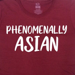 以下が含まれることがあります： ワインレッドのTシャツに、白い文字で「Phenomenally Asian」と書いてあります。