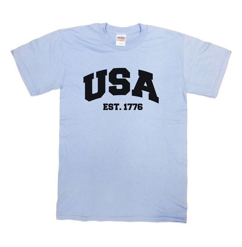 May include: Light blue t-shirt with the word "USA" in bold black letters and "EST. 1776" underneath. The shirt is made of a soft material and has short sleeves and a crew neck.