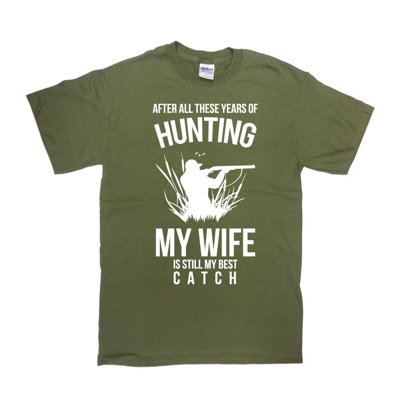 May include: Green t-shirt with white text that reads "After all these years of hunting, my wife is still my best catch". The shirt features a silhouette of a hunter with a rifle.