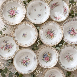 Peut inclure: Ensemble de soupières en porcelaine blanche ornées de motifs floraux. Chaque bol présente un arrangement différent de fleurs roses, bleues et violettes, avec des bordures dorées. Les bols sont disposés sur une surface claire.