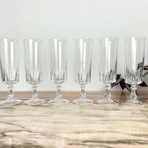 Può includere: Sei flûte da champagne in cristallo trasparente. Ogni bicchiere presenta una coppa alta e sottile con linee incise verticali e una base decorativa. I bicchieri sono esposti su una superficie chiara.