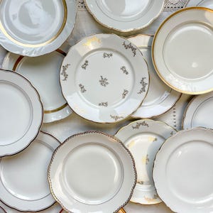 Könnte beinhalten: Eine Sammlung weißer Keramikteller mit goldenen und braunen Verzierungen. Einige Teller haben florale Designs in der Mitte und am Rand. Die Teller variieren in Größe und Form, einige haben einen gewellten Rand.