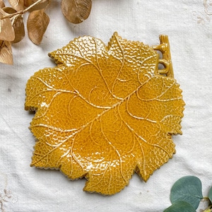 Peut inclure: Un plat décoratif en céramique en forme de feuille, de couleur jaune chaud. Le plat présente des motifs de veines détaillés et une surface texturée, avec une petite poignée en forme de branche. Le design évoque un thème naturel et automnal.