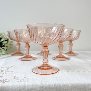 Puede incluir: Un conjunto de seis copas de champán rosas vintage. Cada copa presenta un diseño en espiral en el cuenco y un tallo detallado. Las copas están dispuestas sobre un mantel blanco, mostrando su elegante diseño.