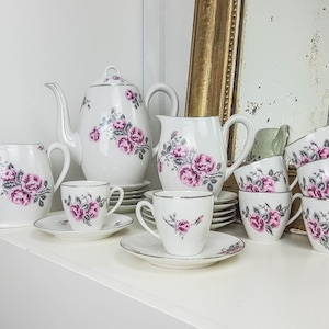 Peut inclure: Un service à thé floral vintage avec une théière, un pot à crème, un sucrier et six tasses et soucoupes. L'ensemble présente un motif floral délicat avec des roses roses et des feuilles grises sur fond blanc.
