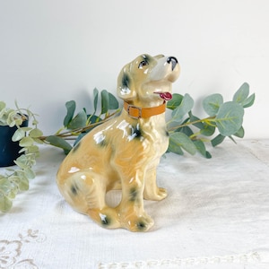 May include: A ceramic dog figurine in a sitting position, with a tan and black spotted coat. The dog has a brown collar and a red tongue. The figurine is a decorative item.