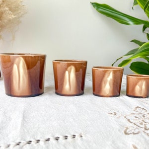 Peut inclure: Quatre pots en céramique brune de tailles différentes avec un glaçage blanc. Les pots sont disposés en rangée du plus grand au plus petit sur une nappe blanche.
