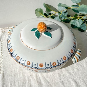 Peut inclure: Plat de service en céramique blanche avec couvercle, orné d'un motif de fruit orange et vert. Le bord est décoré d'un motif floral orange, bleu et blanc. Le plat a des poignées rayées vertes, oranges et blanches.
