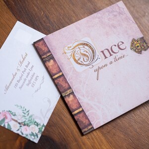 May include: A wedding invitation and envelope set. The invitation features a pink background with the words "Once upon a time..." and a gold emblem. The envelope has floral accents and the names and address of the couple.