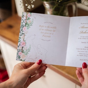 May include: A wedding invitation with floral and fairytale-themed design. The invitation features a floral border with pink and white roses and the text "Right in the middle of an ordinary life love gives us a Fairytale."