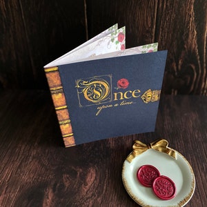May include: A navy blue book with gold lettering that reads "Once upon a time..." and a decorative spine. The book is open, revealing floral patterned pages. Two red wax seals rest on a small, white plate with a gold bow.