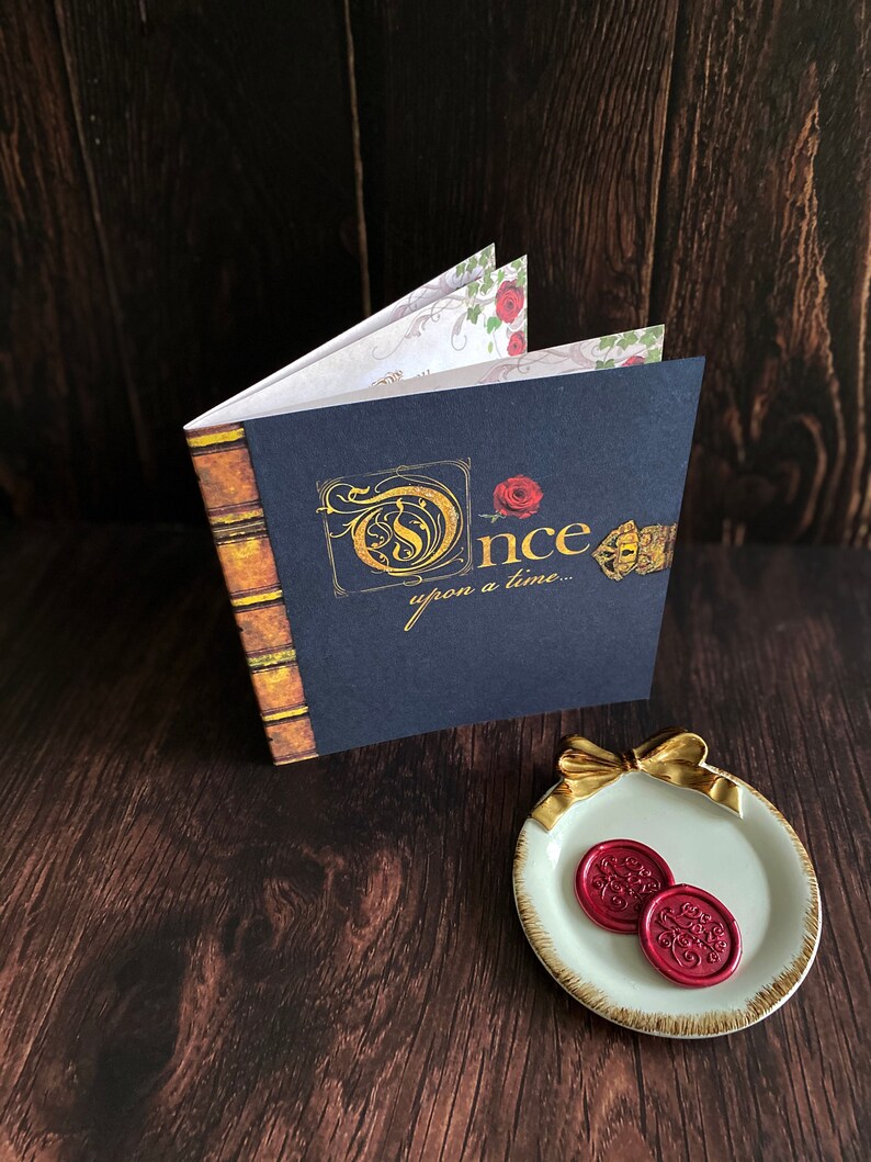 May include: A navy blue book with gold lettering that reads "Once upon a time..." and a decorative spine. The book is open, revealing floral patterned pages. Two red wax seals rest on a small, white plate with a gold bow.