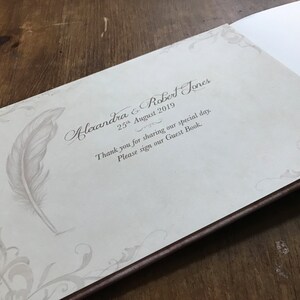 May include: A white guest book with a brown border. The cover features a decorative design with a feather and the text "Alexandra & Robert Jones 25th August 2019 Thank you for sharing our special day. Please sign our Guest Book."