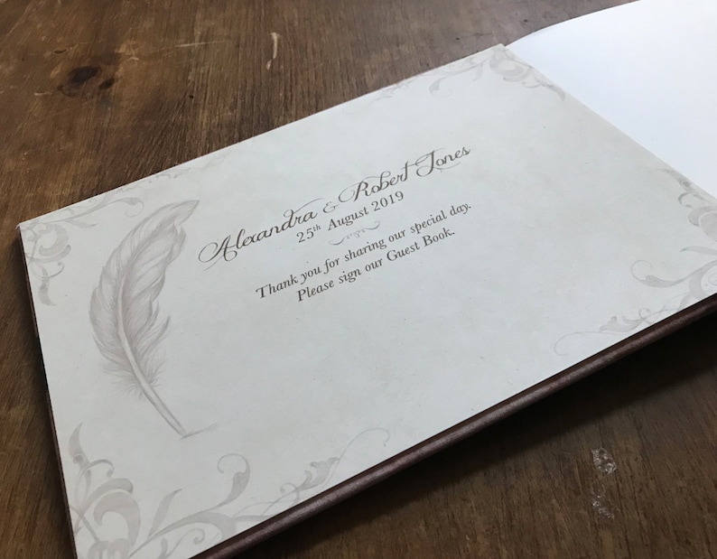 May include: A white guest book with a brown border. The cover features a decorative design with a feather and the text "Alexandra & Robert Jones 25th August 2019 Thank you for sharing our special day. Please sign our Guest Book."