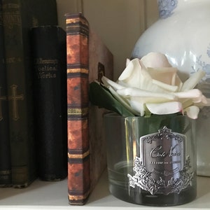 May include: A clear glass jar with a silver label that reads "Cole Noire, L'Elixir de la Beaut&eacute;, Paris" and a white rose inside. The jar is sitting on a shelf with books behind it.