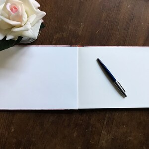 May include: An open blank guest book with a blue pen lying on the right page. The book is on a wooden table with a white rose in a vase in the upper left corner.