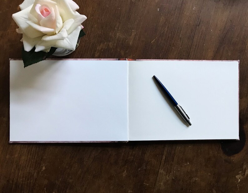May include: An open blank guest book with a blue pen lying on the right page. The book is on a wooden table with a white rose in a vase in the upper left corner.