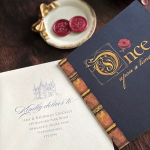 May include: An invitation suite featuring a navy blue booklet with gold lettering that reads "Once upon a time..." and a light cream envelope with a castle illustration and address. Two red wax seals rest on a small white dish.
