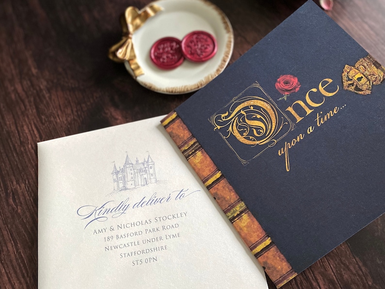 May include: An invitation suite featuring a navy blue booklet with gold lettering that reads "Once upon a time..." and a light cream envelope with a castle illustration and address. Two red wax seals rest on a small white dish.