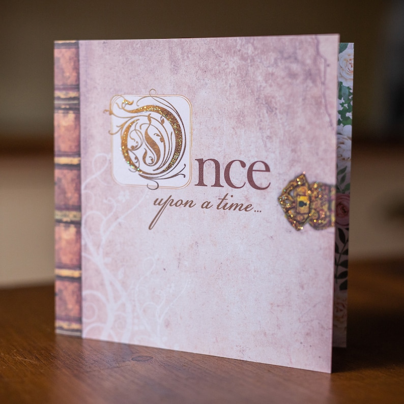 May include: A decorative greeting card with a vintage book design. The cover features the word "Once" and "upon a time..." in a script font, with a gold glitter embellishment. The card has a floral pattern on the inside.