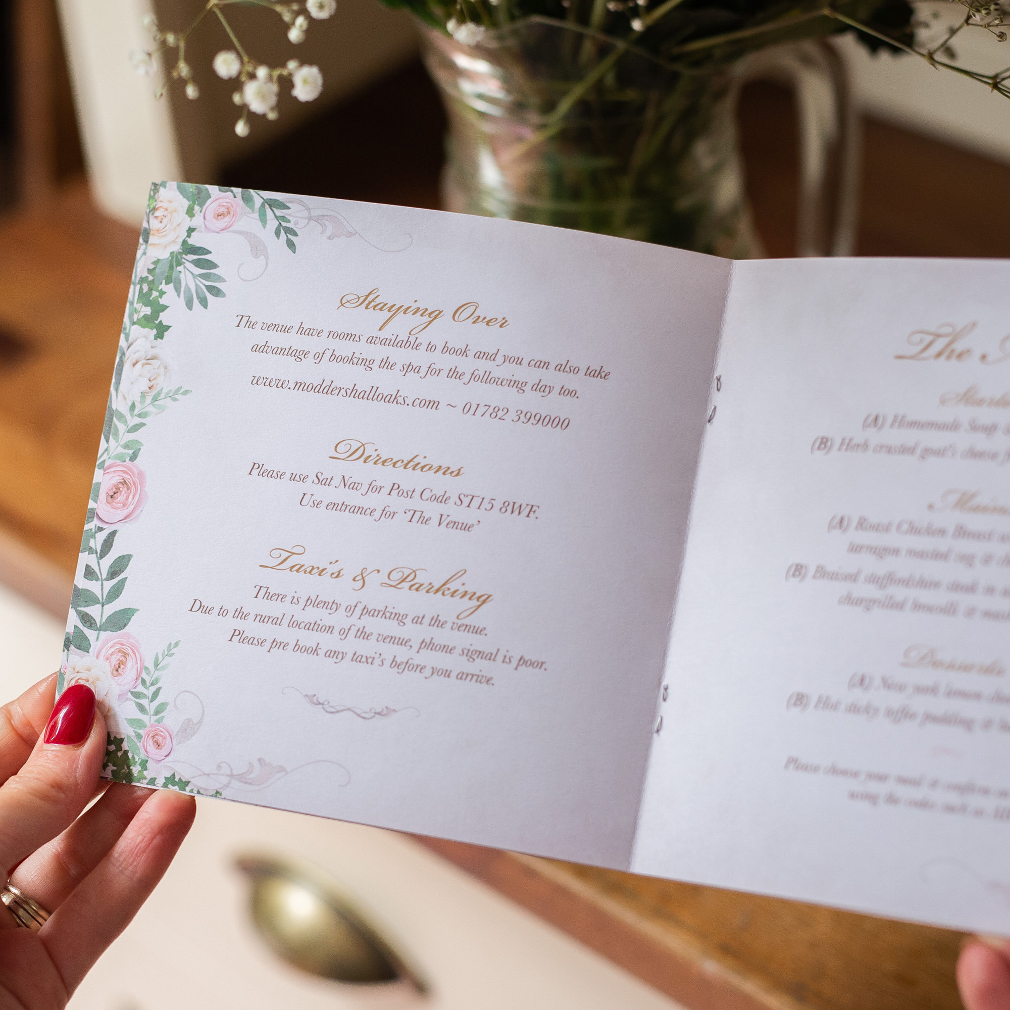 May include: Open wedding invitation with floral and foliage border. The text includes details about staying over, directions, and taxi information. The invitation is held open, revealing the interior details.
