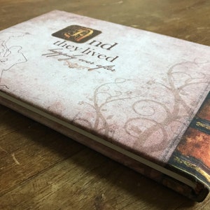 May include: A light brown fabric-covered journal with a decorative spine and the text "And they lived happily ever after." printed on the front cover.