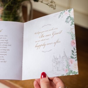 May include: An open wedding invitation with the text "Be our Guest" and "happily ever after" in gold script. The invitation features a castle illustration and floral accents in pink and green.