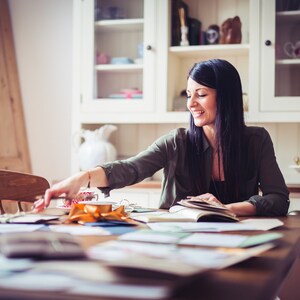May include: A woman with long dark hair smiles while reaching for items on a wooden table. The table is covered with papers, a notebook, and a gold ribbon. A white cabinet with glass doors is in the background.