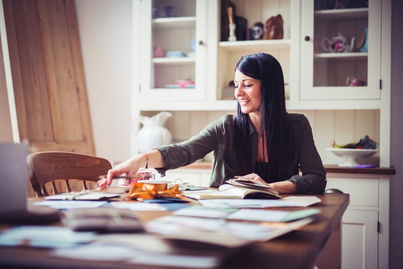 May include: A woman with long dark hair smiles while reaching for items on a wooden table. The table is covered with papers, a notebook, and a gold ribbon. A white cabinet with glass doors is in the background.
