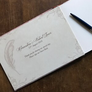 May include: A white guest book with a decorative floral design and a gray feather pen illustration. The text on the cover reads "Alexandra & Robert Jones 25th August 2019 Thank you for sharing our special day. Please sign our Guest Book."