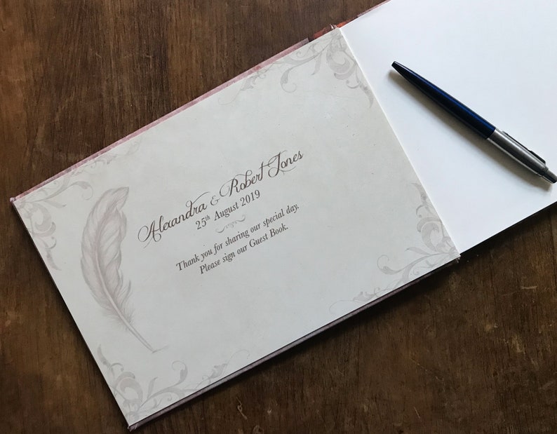 May include: A white guest book with a decorative floral design and a gray feather pen illustration. The text on the cover reads "Alexandra & Robert Jones 25th August 2019 Thank you for sharing our special day. Please sign our Guest Book."