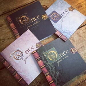 May include: Four square-shaped journals with the words "Once upon a time..." in elegant script. Each journal has a different color scheme, including black, blue, gray, and pink, with gold accents and a decorative spine.