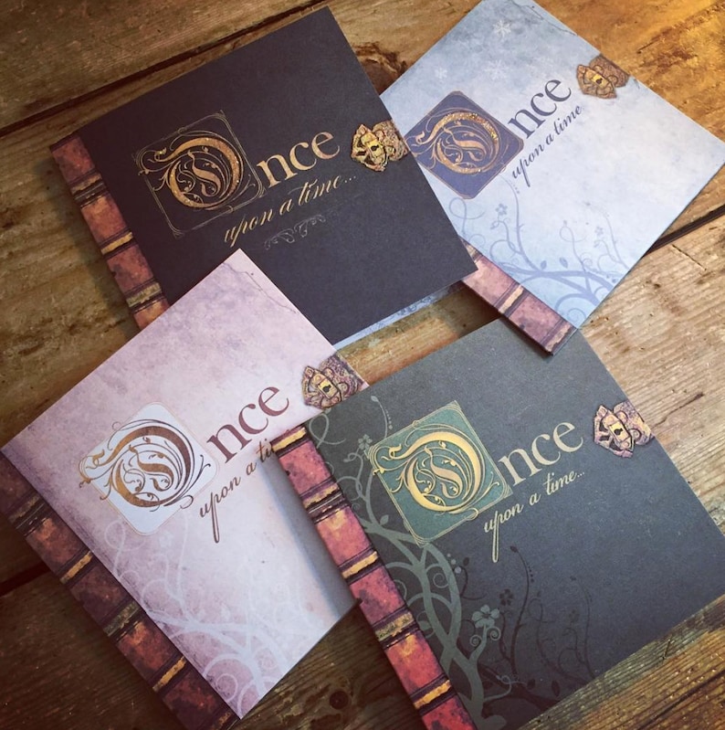 May include: Four square-shaped journals with the words "Once upon a time..." in elegant script. Each journal has a different color scheme, including black, blue, gray, and pink, with gold accents and a decorative spine.