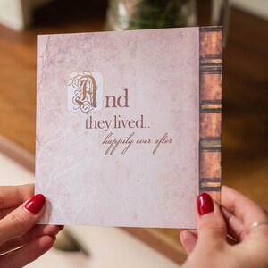 May include: A square card with a vintage design, held by a person. The card features the text "And they lived... happily ever after" in a script font. The left side has a faded pink background with a decorative flourish, and the right side has a faux book spine.